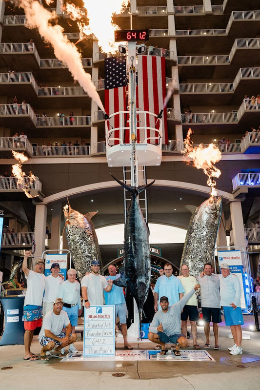A Work of Art's crew poses with the 641.2-pound winning blue marlin hanging at The Wharf's Marlin Circle scale as fire cannons erupt at the 2026 Blue Marlin Grand Championship