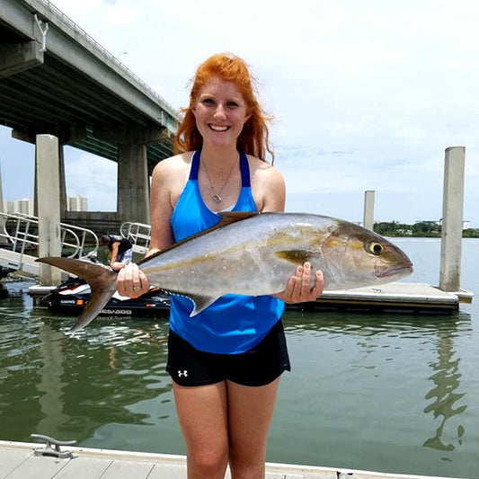 A charter angler holds a greater amberjack at a Daytona Beach dock, a bridge spanning the intracoastal behind her.