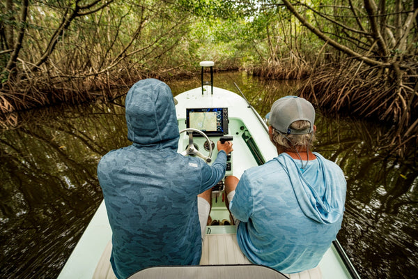 Two anglers wearing Huk A1A Pro sun hoodies run a flats skiff through a narrow mangrove creek